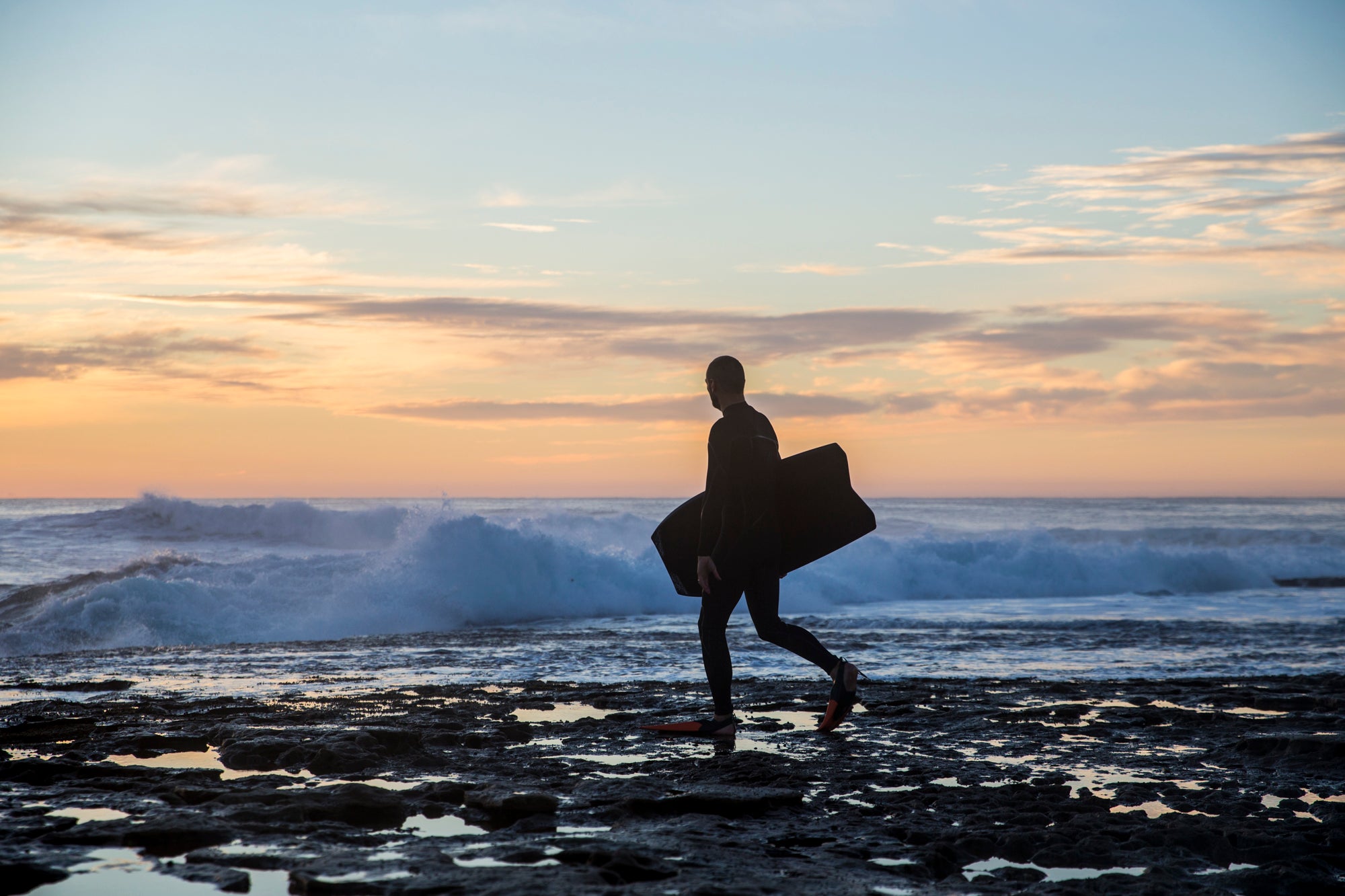 A man holds a bodyboard as he approaches the ocean, a wave breaks in the background and there is a pretty sunset backdrop. 